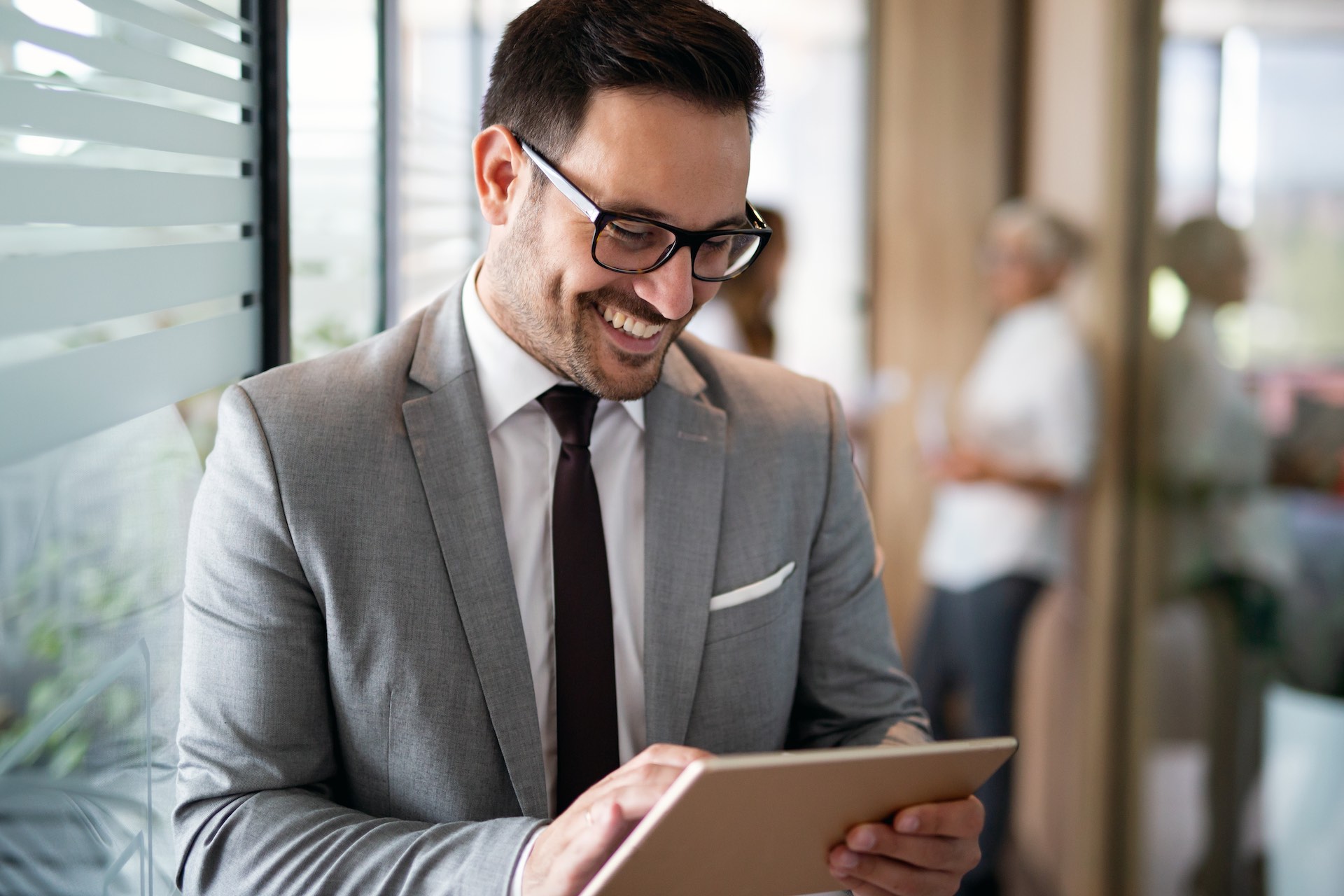 Happy young business man using a digital tablet while standing in front of windows in office