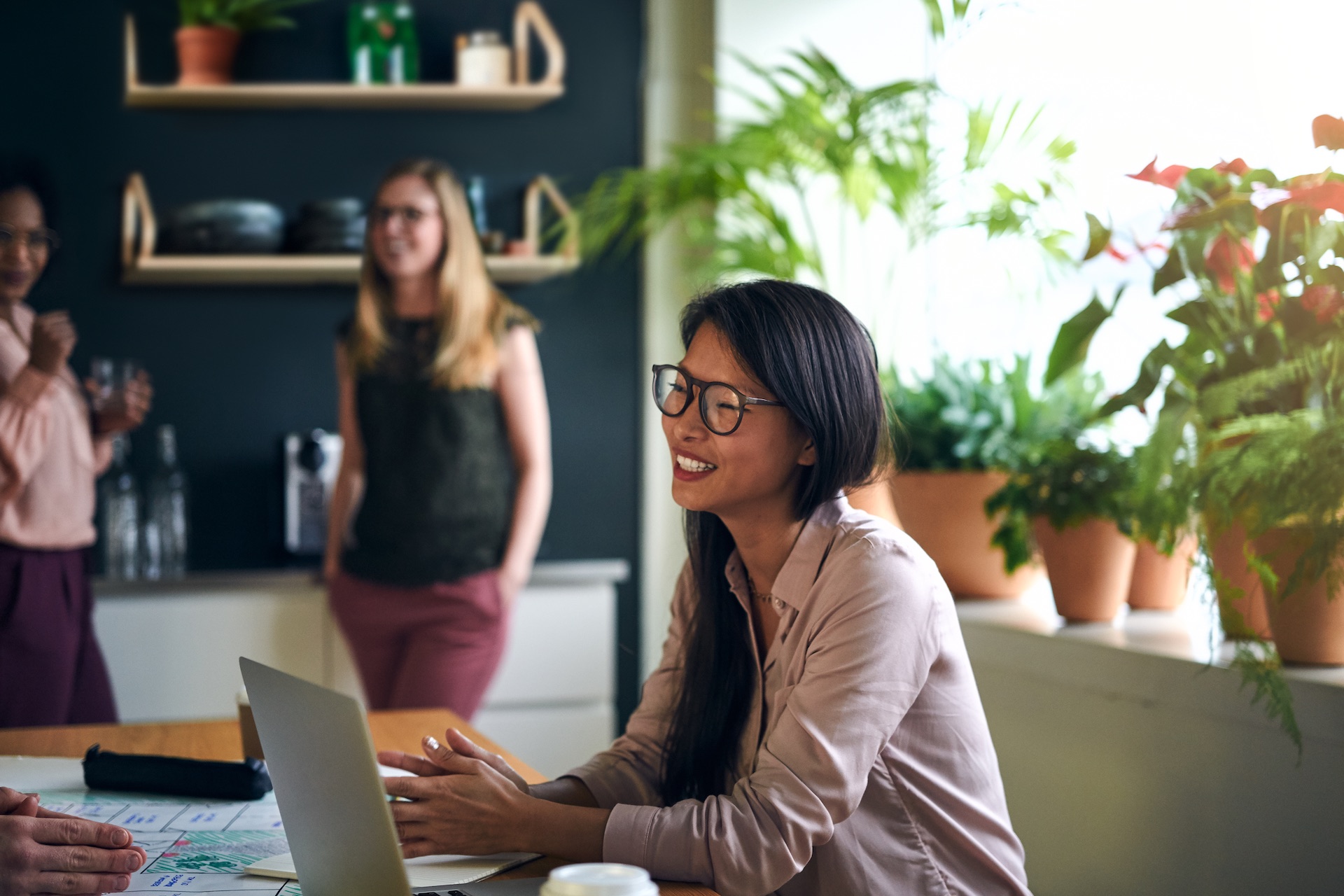 Smiling Asian businesswoman sitting at a table in a modern office using a laptop with colleagues working in the background