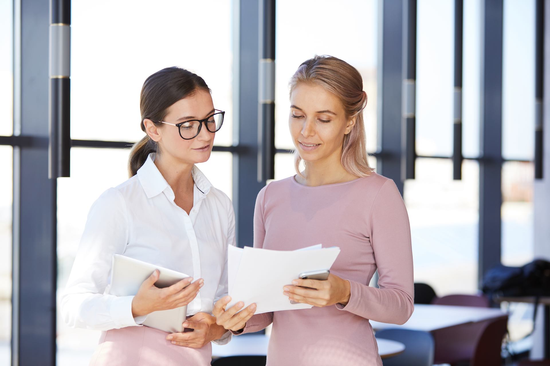 Content confident young businesswomen standing in lobby and viewing papers together while planning product promotion