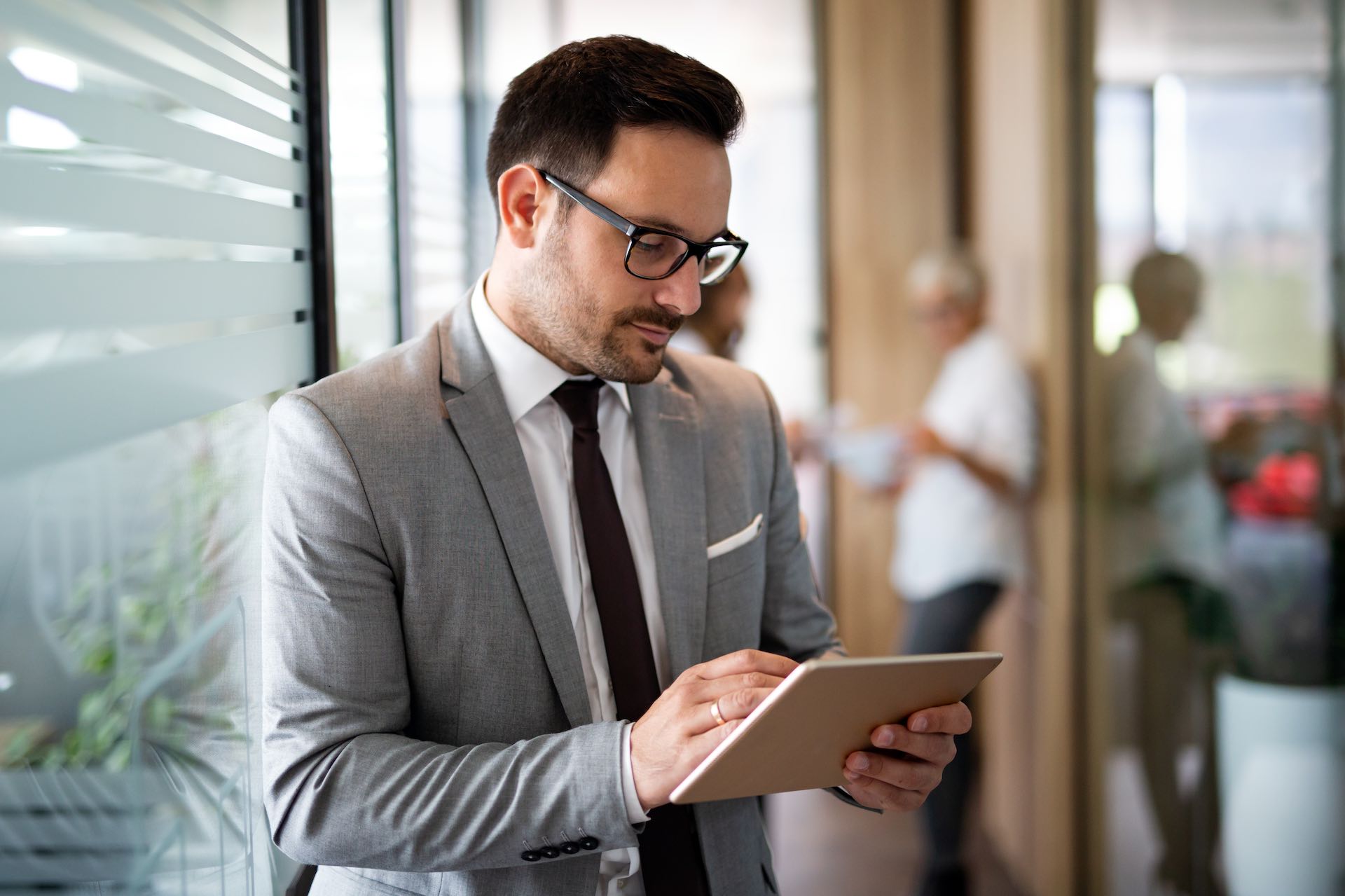 Happy young business man using a digital tablet while standing in front of windows in office