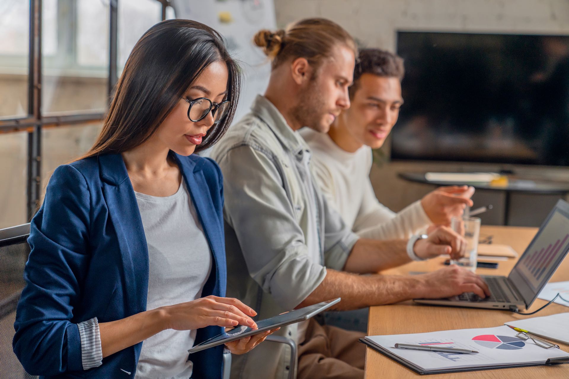 Asian female working on digital tablet. Young businessman in meeting with coworkers in conference room. Creative professionals discussing business in board room