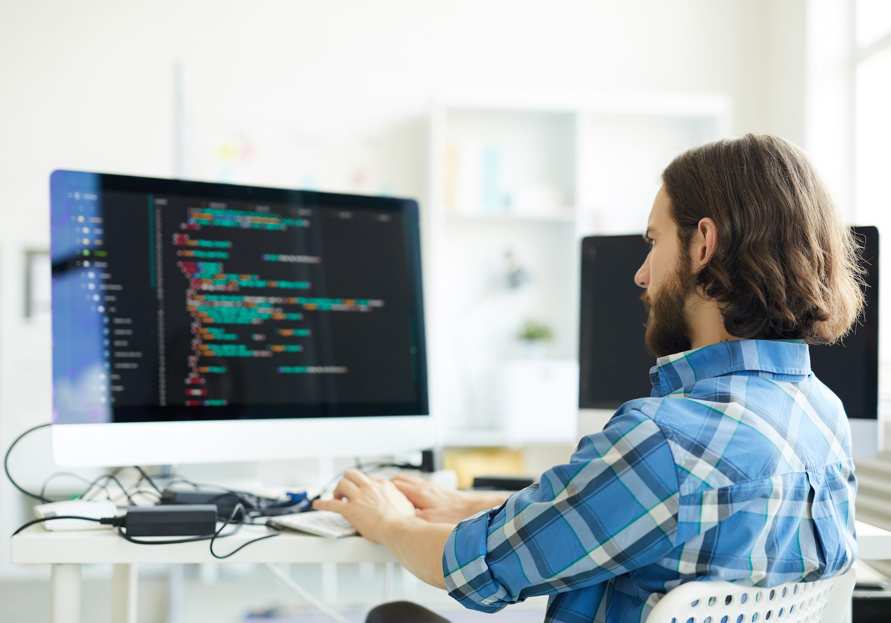 Serious pensive hipster young bearded coder in casual shirt creating computer software and working with desktop computer in modern office