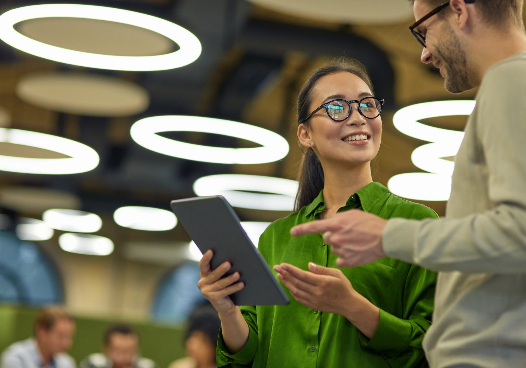 Discussing project. Young positive asian woman showing something on digital tablet to her male colleague while standing together in the coworking space or office. Teamwork, business people at work